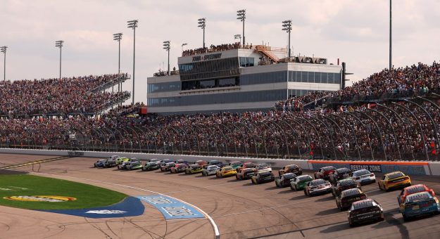 General view of packed stands at Iowa Speedway as cars come past the start-finish line.